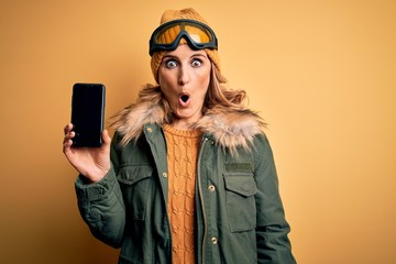 Middle age blonde skier woman wearing ski goggles holding smartphone showing screen scared in shock...
