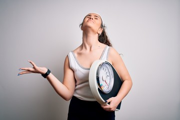Young beautiful woman with curly hair holding weighing machine over white background crazy and mad...