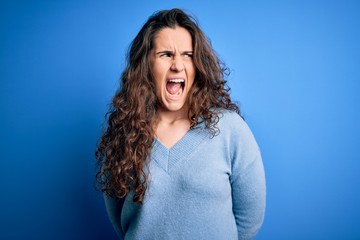 Young beautiful woman with curly hair wearing blue casual sweater over isolated background angry and mad screaming frustrated and furious, shouting with anger. Rage and aggressive concept.