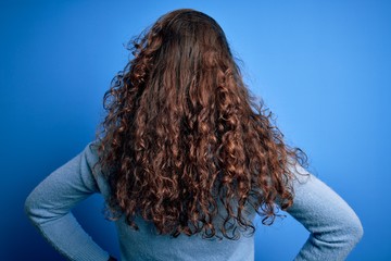 Young beautiful woman with curly hair wearing blue casual sweater over isolated background standing backwards looking away with arms on body