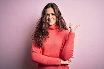 Fototapeta premium Young beautiful woman with curly hair wearing turtleneck sweater over pink background smiling with happy face looking and pointing to the side with thumb up.