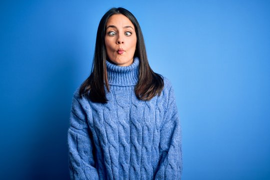 Young brunette woman with blue eyes wearing casual turtleneck sweater making fish face with lips, crazy and comical gesture. Funny expression.