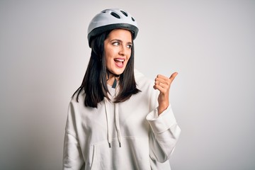 Young cyclist woman with blue eyes wearing bike helmet over isolated white background smiling with happy face looking and pointing to the side with thumb up.