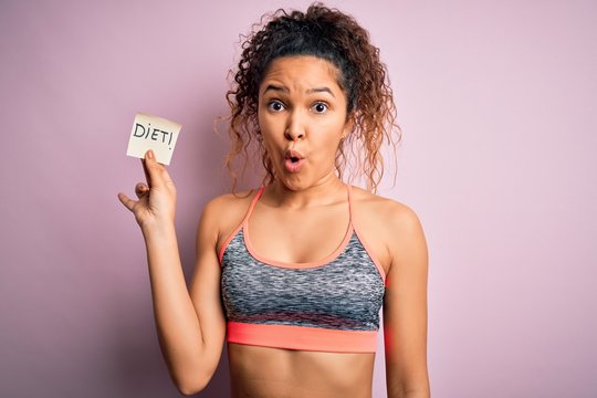 Beautiful sportswoman with curly hair doing sport wearing sportswear holding diet reminder scared in shock with a surprise face, afraid and excited with fear expression