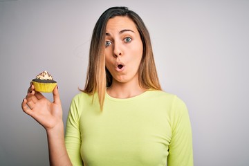 Young beautiful woman with blue eyes eating sweet chocolate cupcake over white background scared in shock with a surprise face, afraid and excited with fear expression