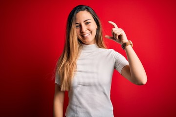 Fototapeta premium Beautiful blonde woman with blue eyes wearing casual white t-shirt over red background smiling and confident gesturing with hand doing small size sign with fingers looking and the camera. Measure 