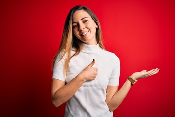 Obraz premium Beautiful blonde woman with blue eyes wearing casual white t-shirt over red background Showing palm hand and doing ok gesture with thumbs up, smiling happy and cheerful