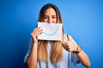 Young beautiful brunette woman holding card with smile illustration on mouth happy with big smile doing ok sign, thumb up with fingers, excellent sign