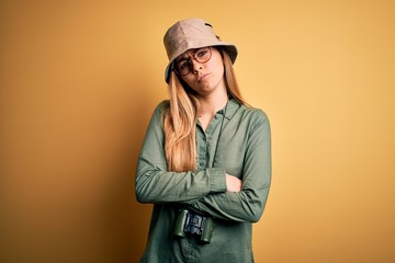 Beautiful blonde explorer woman with blue eyes wearing hat and glasses using binoculars skeptic and...