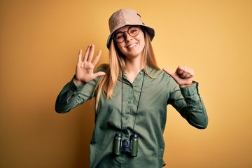 Beautiful blonde explorer woman with blue eyes wearing hat and glasses using binoculars showing and pointing up with fingers number six while smiling confident and happy.