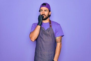 Young handsome hispanic man with bear wearing professional apron working as tattoo artist looking stressed and nervous with hands on mouth biting nails. Anxiety problem.