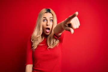 Young beautiful blonde woman wearing casual t-shirt standing over isolated red background Pointing with finger surprised ahead, open mouth amazed expression, something on the front