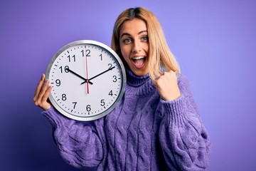 Young beautiful blonde woman doing countdown holding big clock over purple background screaming proud and celebrating victory and success very excited, cheering emotion