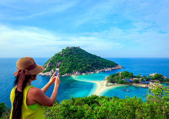 Fototapeta premium Female tourist using smartphone taking picture of beautiful landscape of islands and blue sea with tropical white sand beach in Thailand