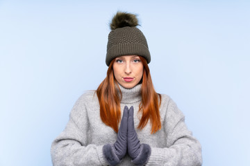 Young redhead Russian woman with winter hat over isolated blue background pleading