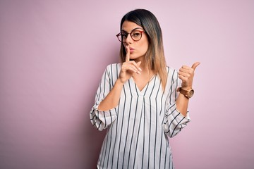 Young beautiful woman wearing casual striped t-shirt and glasses over pink background asking to be quiet with finger on lips pointing with hand to the side. Silence and secret concept.
