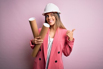 Young beautiful brunette architect woman wearing safety helmet holding blueprints pointing and showing with thumb up to the side with happy face smiling