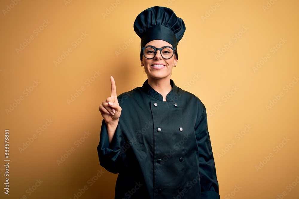 Canvas Prints Young beautiful brunette chef woman wearing cooker uniform and hat over yellow background showing and pointing up with finger number one while smiling confident and happy.