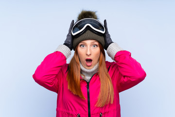 Skier redhead woman with snowboarding glasses over isolated blue wall with surprise facial expression