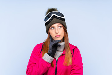 Skier redhead woman with snowboarding glasses over isolated blue wall thinking an idea