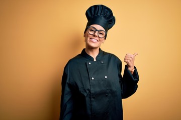 Young beautiful brunette chef woman wearing cooker uniform and hat over yellow background smiling...