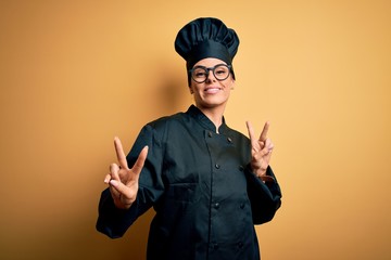 Young beautiful brunette chef woman wearing cooker uniform and hat over yellow background smiling looking to the camera showing fingers doing victory sign. Number two.