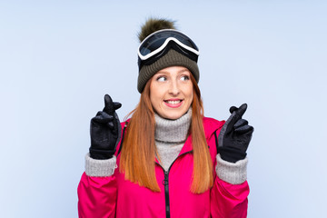 Skier redhead woman with snowboarding glasses over isolated blue wall with fingers crossing