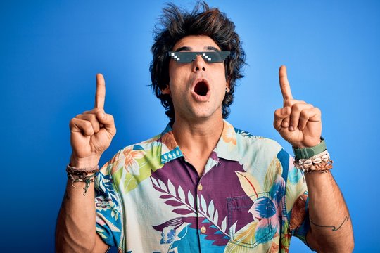 Young handsome man wearing thug life glasses standing over isolated blue background amazed and surprised looking up and pointing with fingers and raised arms.