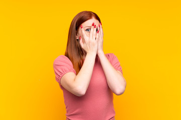 Young redhead woman over isolated yellow background covering eyes and looking through fingers