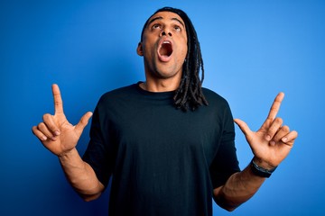 Young handsome african american afro man with dreadlocks wearing casual t-shirt amazed and surprised looking up and pointing with fingers and raised arms.