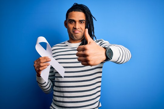 Young african american man with dreadlocks holding white ribbon over blue background happy with big smile doing ok sign, thumb up with fingers, excellent sign