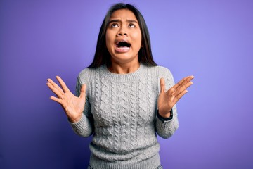 Young beautiful asian girl wearing casual sweater standing over isolated purple background crazy and mad shouting and yelling with aggressive expression and arms raised. Frustration concept.