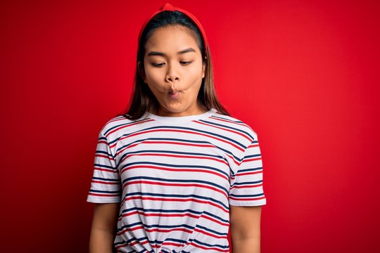 Young beautiful asian girl wearing casual striped t-shirt over isolated red background making fish face with lips, crazy and comical gesture. Funny expression.