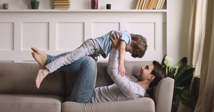 Full length side view strong young father lifting on arms small playful kid son, having fun together on couch at home. Happy two male generations family practicing acroyoga or playing on sofa.