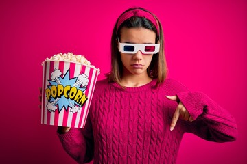 Obraz premium Young beautiful girl watching movie using 3d glasses eating box with popcorns Pointing down looking sad and upset, indicating direction with fingers, unhappy and depressed.