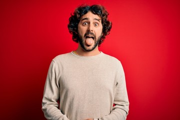 Fototapeta na wymiar Young handsome man with beard wearing casual sweater standing over red background sticking tongue out happy with funny expression. Emotion concept.