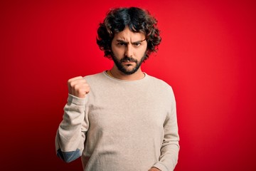 Young handsome man with beard wearing casual sweater standing over red background angry and mad...