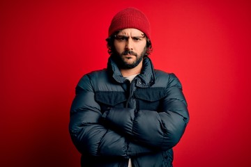 Obraz premium Young handsome man with beard wearing casual coat and wool cap over red background skeptic and nervous, disapproving expression on face with crossed arms. Negative person.