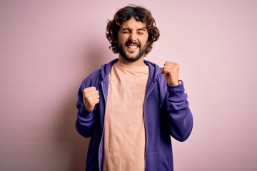 Young handsome sporty man with beard wearing casual sweatshirt over pink background celebrating surprised and amazed for success with arms raised and eyes closed. Winner concept.