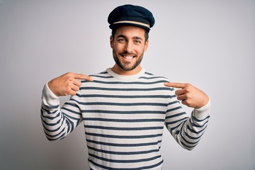 Fototapeta premium Young handsome sailor man with beard wearing navy striped uniform and captain hat looking confident with smile on face, pointing oneself with fingers proud and happy.