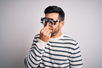 Young handsome man with beard wearing optometry glasses over isolated white background smelling something stinky and disgusting, intolerable smell, holding breath with fingers on nose. Bad smell