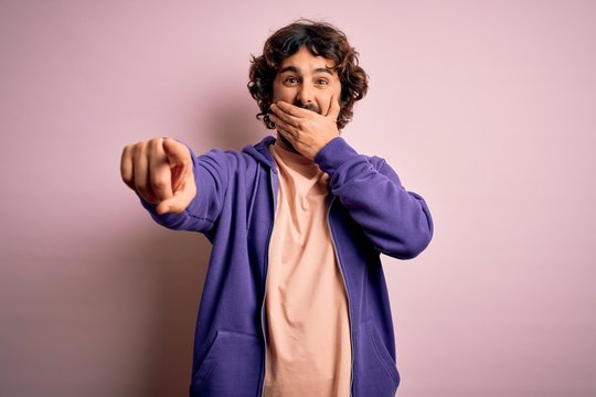Young handsome sporty man with beard wearing casual sweatshirt over pink background laughing at you, pointing finger to the camera with hand over mouth, shame expression