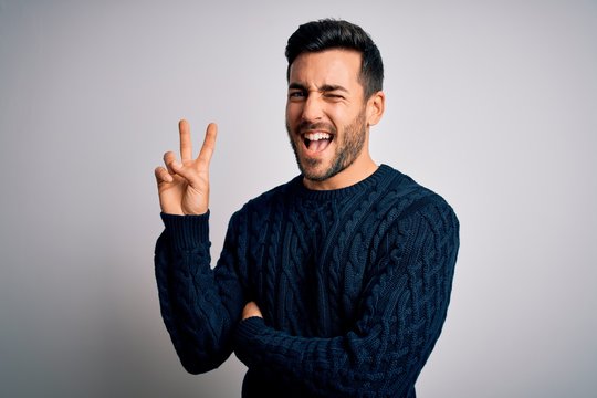 Young handsome man with beard wearing casual sweater standing over white background smiling with happy face winking at the camera doing victory sign. Number two.