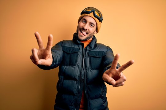 Young handsome skier man with beard wearing snow sportswear and ski goggles smiling with tongue out showing fingers of both hands doing victory sign. Number two.