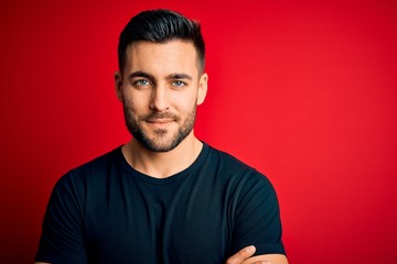 Obraz premium Young handsome man wearing casual black t-shirt standing over isolated red background happy face smiling with crossed arms looking at the camera. Positive person.