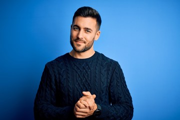 Young handsome man wearing casual sweater standing over isolated blue background with hands together and crossed fingers smiling relaxed and cheerful. Success and optimistic