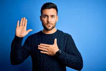 Young handsome man wearing casual sweater standing over isolated blue background Swearing with hand...