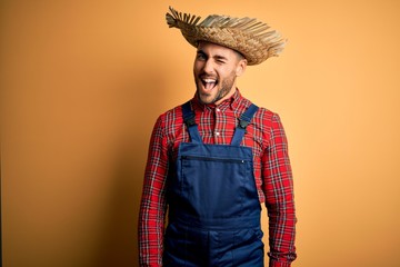 Young rural farmer man wearing bib overall and countryside hat over yellow background winking...