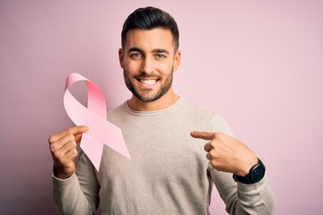 Young handsome man holding pink cancer ribbon symbol of support over isolated background with...
