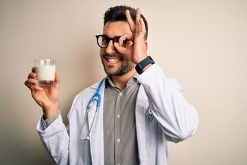Young doctor man wearing stethoscope holding a glass of milk over isolated background with happy face smiling doing ok sign with hand on eye looking through fingers